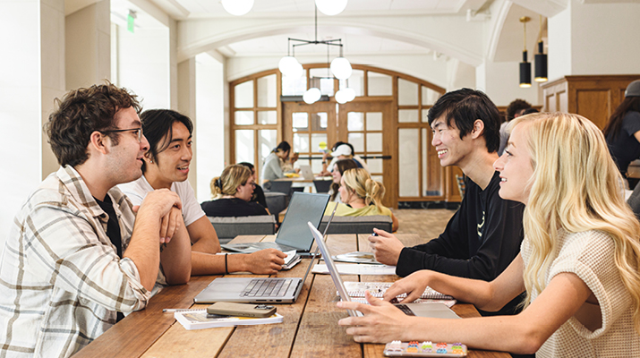 Four students sitting at wood table with laptops facing each other